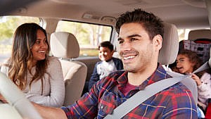 A man smiles as he drives a car with his wife and family