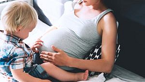 A pregnant mom sitting on a bed. Their child is sitting on her lap, facing mom, and is touching her stomach.