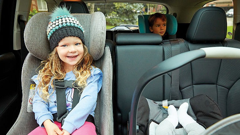 a little girl smiles in the car in her convertible car seat next to a baby in a bucket seat and with a big kid in a booster in the third row