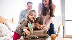 Photo of two kids helping their mom close a suitcase while dad watches