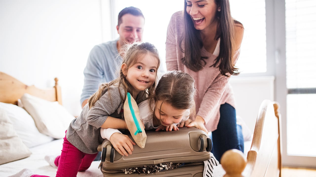 Photo of two kids helping their mom close a suitcase while dad watches