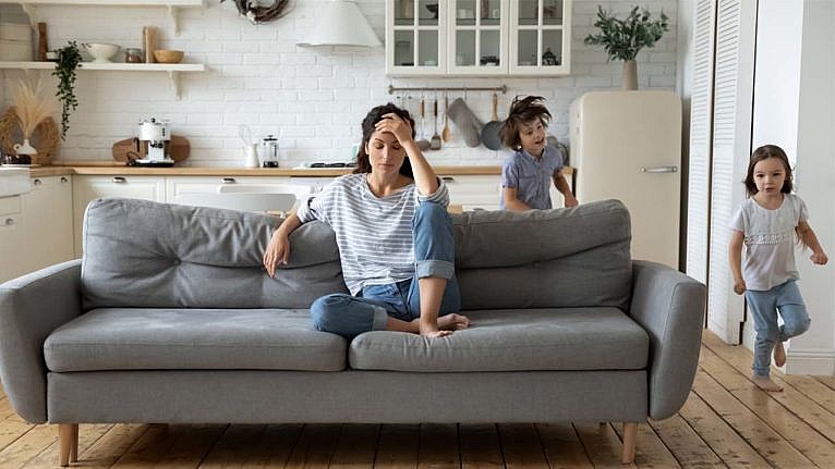 Parent sitting on a grey couch stressed. There two children are running around their parent.
