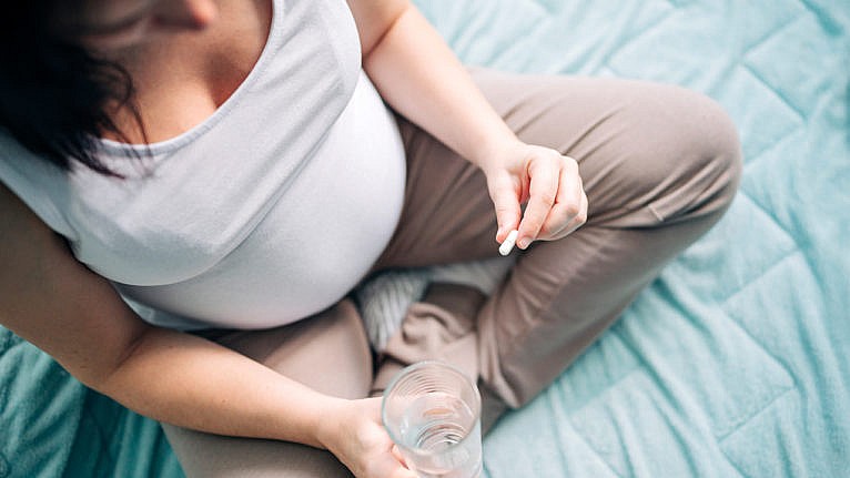 a pregnant woman sits on her bed cross-legged with a pill and a cup of water