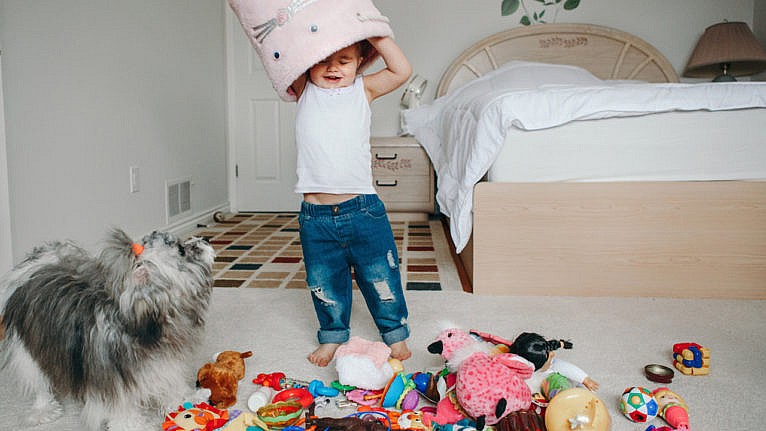 a toddler who dumped out her toys and put the pink bin on her head while her dog watches