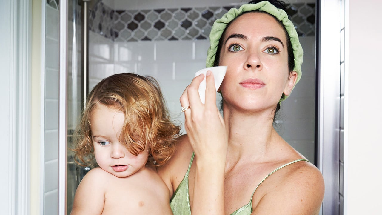a mom with her hair back in a headband wipes her face down with toner while holding her toddler