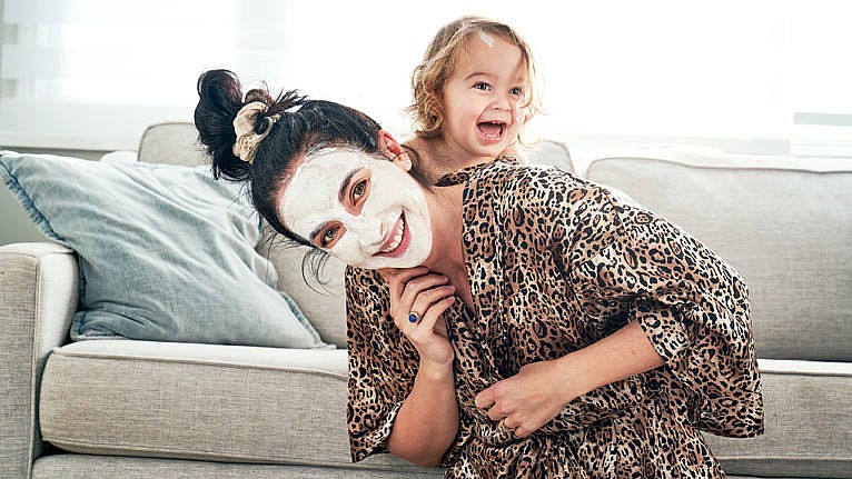 A woman wearing a white clay mask smiles with her baby laughing behind her on the sofa.