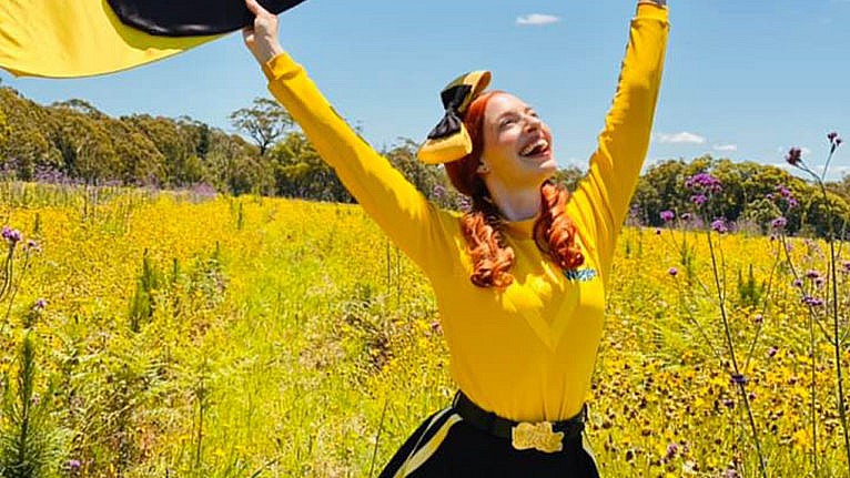 Photo of Emma dressed as the Yellow Wiggle standing in a field holidng up a big farbic version of her yellow bow.