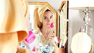 A woman wearing a towel over her head looking in the mirror while rolling a pink face roller on her cheek.