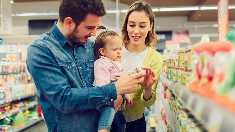 A mother and father hold their toddler. They're standing in a grocery store aisle looking at a food label.