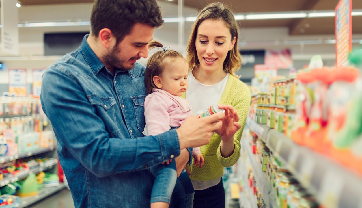 A mother and father hold their toddler. They're standing in a grocery store aisle looking at a food label.