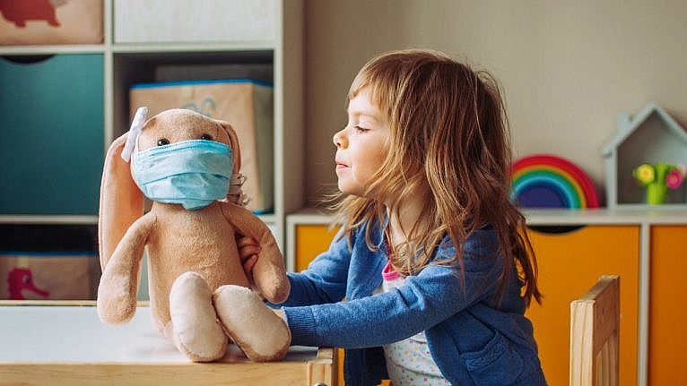A young child sits in a play room. She is holding her stuffed bunny on the table next to her. The bunny is wearing a blue surgical mask.
