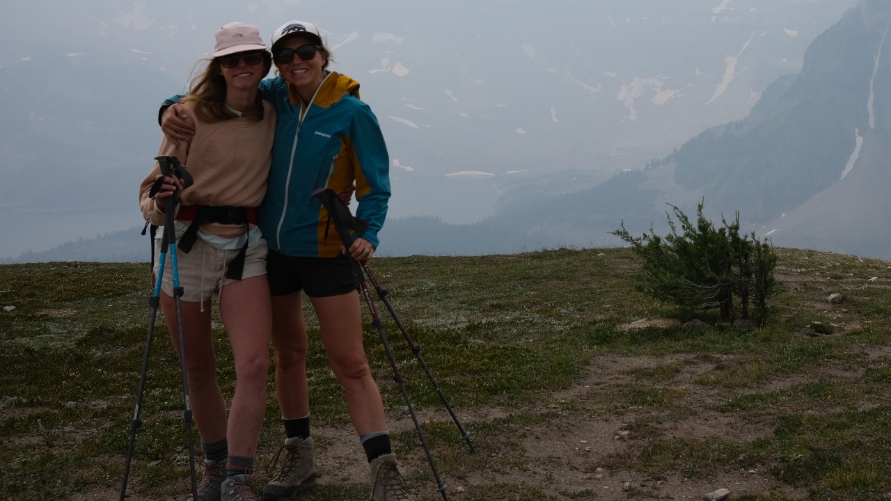 Two women standing next to each other on a hill in front of mountains.