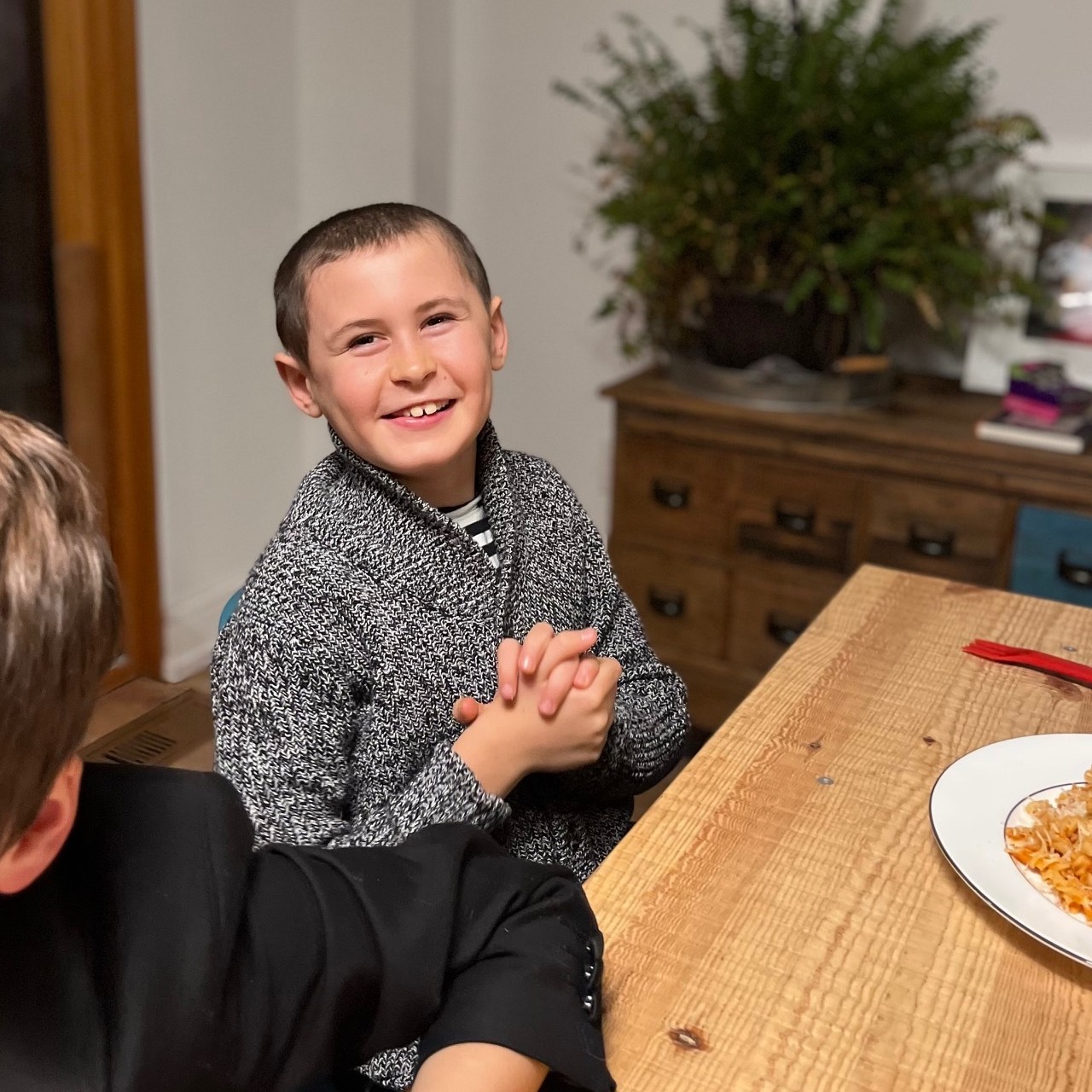 A smiling boy sits at a table with a plate of pasta in front of him.