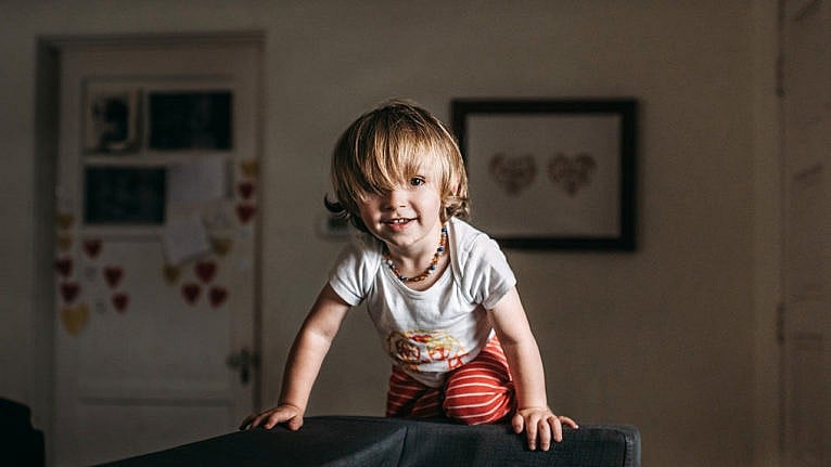 A toddler smiling while climbing on the arm of a couch.
