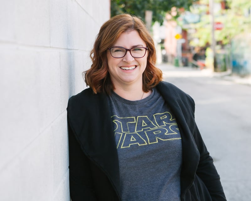 A woman with red hair and glasses smiles while leaning against a white brick wall. She is wearing a "Star Wars" shirt and a blazer.