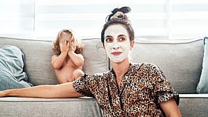 A woman leans against the side of her couch. She's wearing a cheetah print silk pyjama shirt and a white face mask. Her kid sits on the couch behind her.