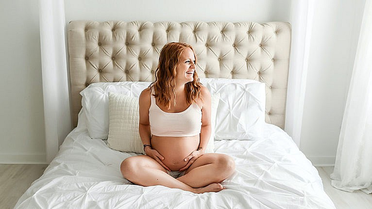 Redheaded pregnant woman sits cross-legged on her white bed holding her belly.