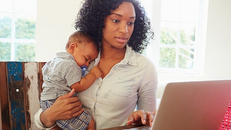 A woman does work at her laptop while holding her baby.