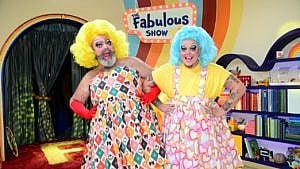 An image of drag duo Fay and Fluffy; Fay is wearing a yellow wig with a patterned dress and red gloves; Fluffy is wearing a blue wig with a yellow t-shirt and a pink heart dress.