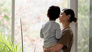 A woman holds her son, who appears to be a toddler. She is standing near a bright window and looking at him in a sad way.