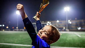 A young boy wearing a blue soccer jersey smiling and holding up a golden trophy.