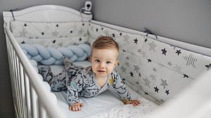 An image of an unhappy baby in a white crib with star-patterned crib bumpers around him.
