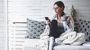 A woman sitting on the couch looking at her phone while holding a mug.