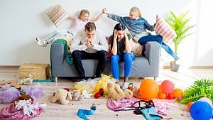 Exasperated parents sit on a couch with their heads in their hands while their two daughters high five behind them. The house around them is messy.