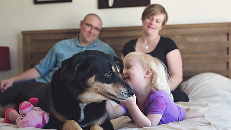 A mother and father sit on their bed watching while their daughter cuddles the family dog