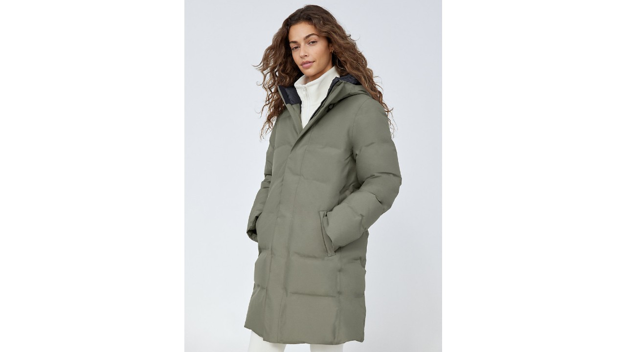 A woman wears a green parka. She has her hands in its pockets.