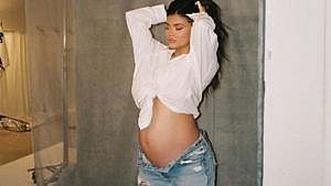 Kylie Jenner stands in front of a grey wall. She's holding her hair up and her pregnant belly is exposed.