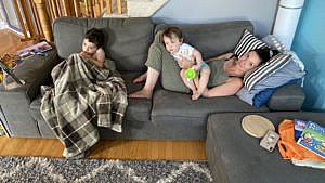 A mom lays on the couch with her two kids. One of the kids is covered in a blanket while the other sits on mom's belly.