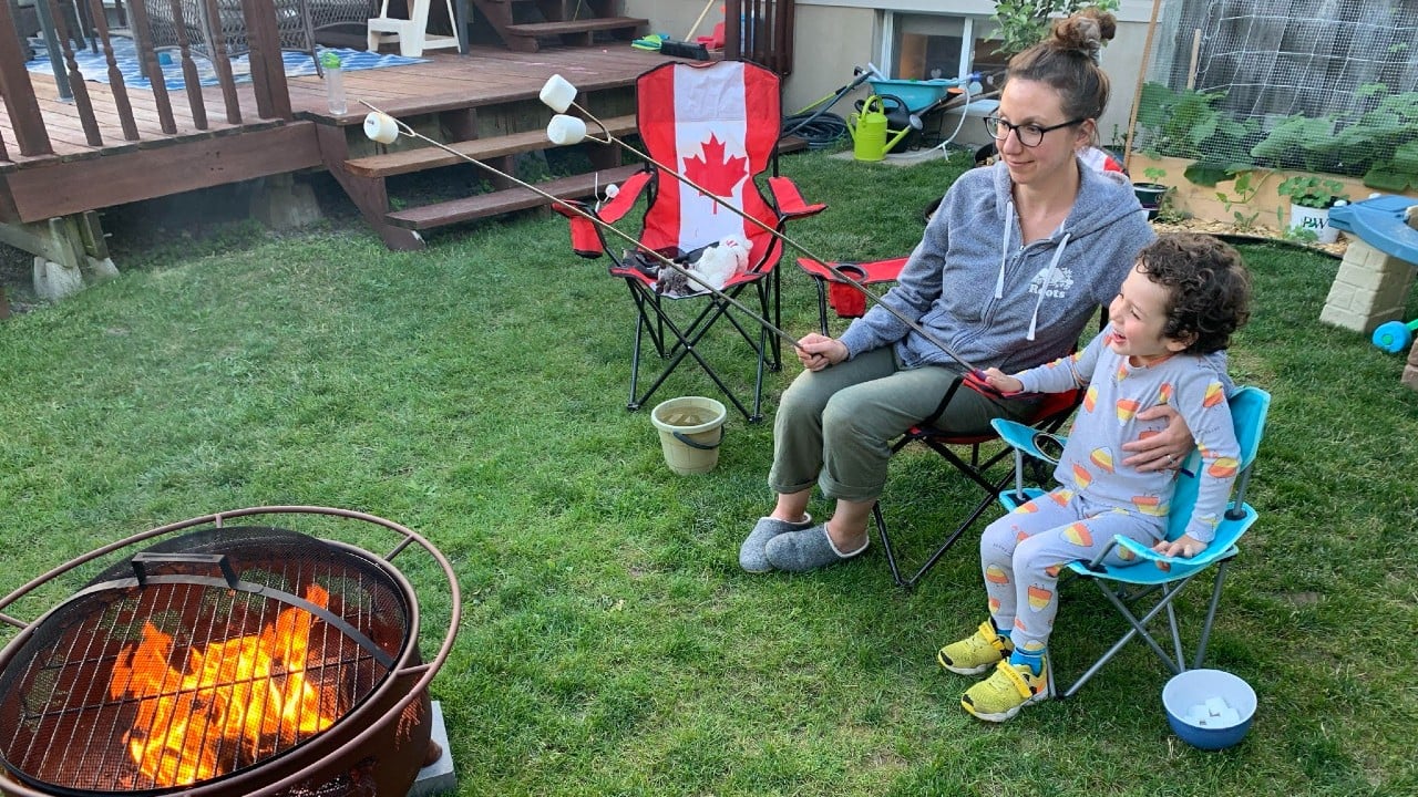 A mom and her young kid sit on fold-up camping chairs in her backyard. They're holding big skewers with marshmallows on them over a fire pit