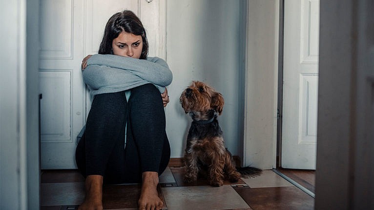 A sad woman sit on the floor with her knees pulled into her chest and her dog beside her