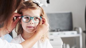 A little girl in a white t-shirt sits while a woman places bright blue eye glasses on her face