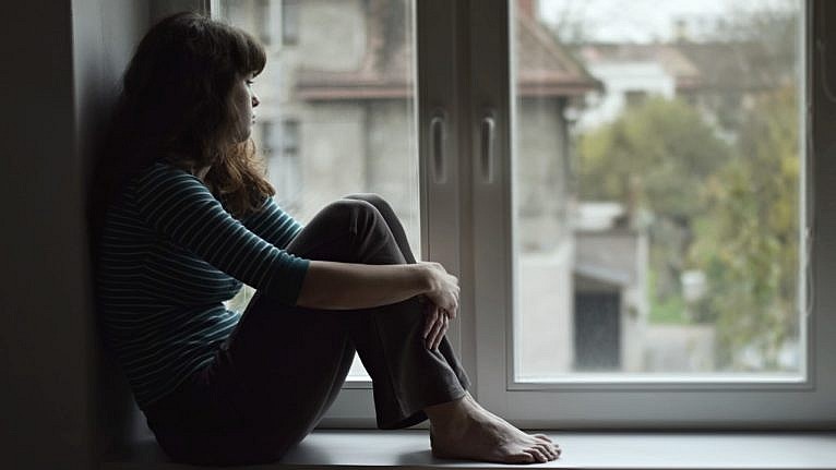 An image of a woman sitting on a windowsill looking out a window.