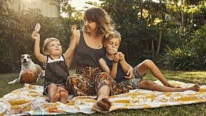A mother sitting with her two kids outside on a picnic blanket with their dog.