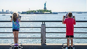 Two kids look through coin-operated binocular stations out at the statue of liberty