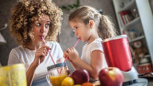 A woman and her daughter drinking from a smoothie cup with straws, with a blender and fruit on the table. The mother looks at her daughter's face intently for her reaction.