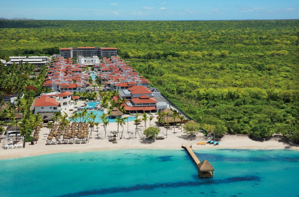 Dreams Dominicus La Romana Resort is seen from above, with a beach with lounge chairs, a pier in the water, and hotel buildings visible