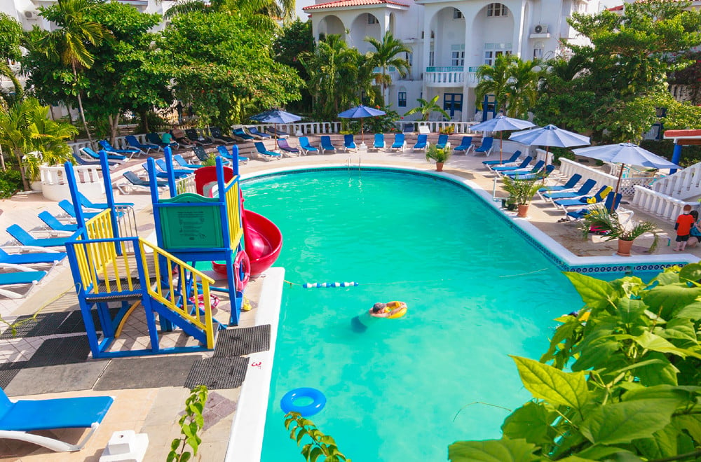 A pool with a kids' playset, with lounge chairs and a hotel building at Franklyn D. Resort in Jamaica