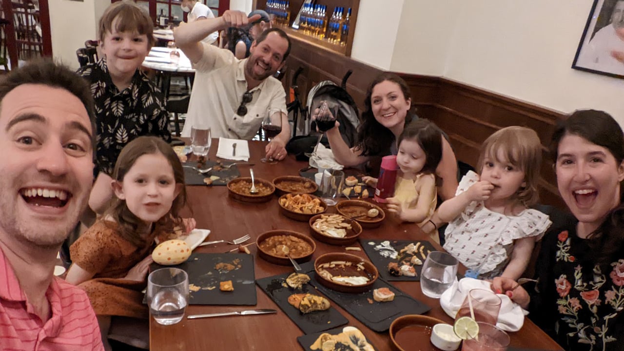 Two families with four kids between them smile at the camera sitting around a table with lots of tapas foods on it