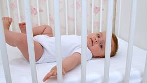 A four-month-old baby wearing a white t-shirt onesie lies in their crib wide awake