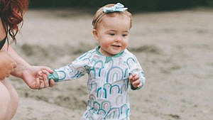 A toddler on the beech wears a long-sleeved bathing suit and matching headband