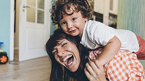 A mom laughs heartily while playing with her toddler, who is climbing on her back