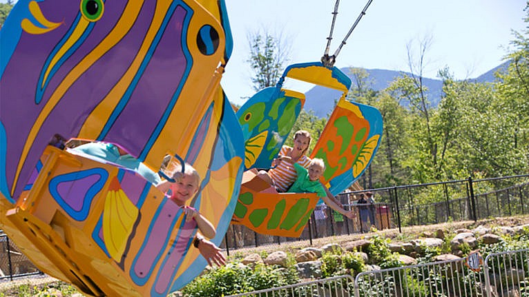 A family rides in a swinging fish-themed ride with mountains in the background