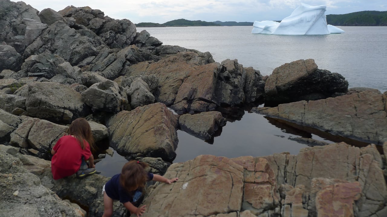 Two kids playing in rock pools as an iceberg floats by in the distance.