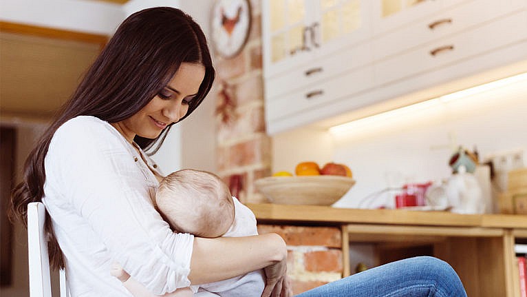 A mom nurses her baby while sitting in her kitchen
