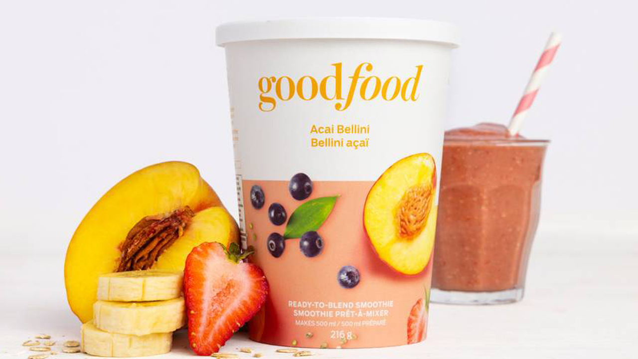 A ready-to-blend MadeGood smoothie cup beside a half peach, strawberry and stack of banana slices with a blended smoothie and striped straw in the background