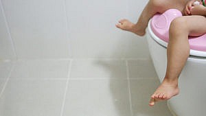 A small child's legs dangle off the edge of a toilet seat fitted with a potty attachment.